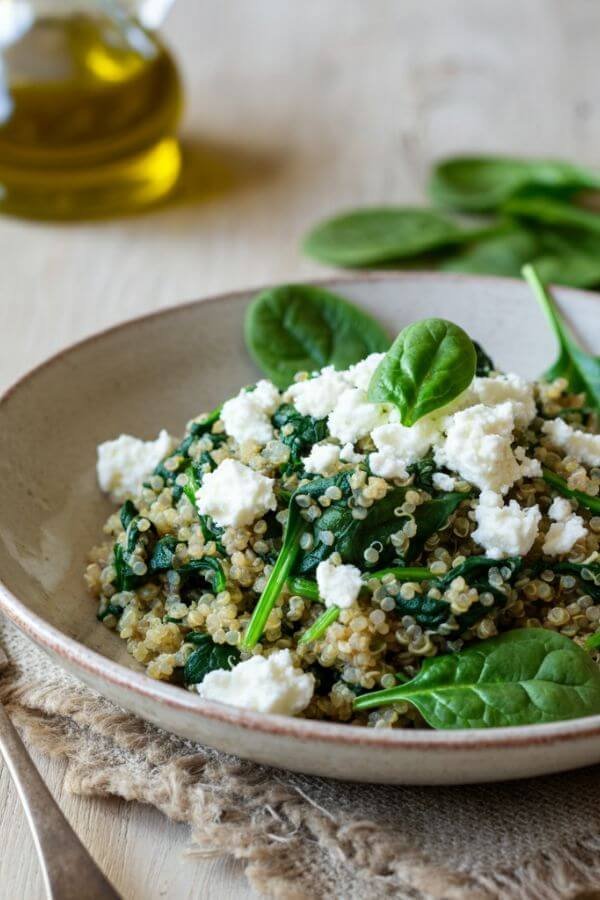 A close look of A white bowl of vibrant quinoa topped with sautéed spinach and crumbled feta cheese, drizzled with olive oil. The dish is garnished with a few spinach leaves on the side, placed on a light wooden table with a rustic fork nearby.