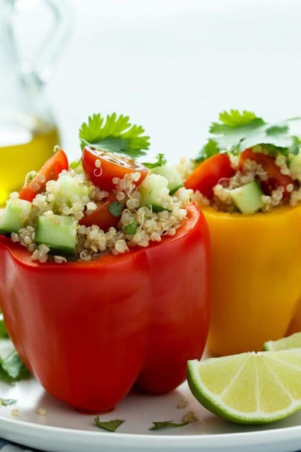 A vibrant image of two stuffed bell peppers, one red and one yellow, placed on a clean white plate. The bell peppers are filled with a colorful mix of sprouted quinoa, diced cucumbers, halved cherry tomatoes, and fresh cilantro, with lime wedges and a drizzle of olive oil on the side. The background is bright and minimal, with natural lighting highlighting the textures and fresh ingredients.