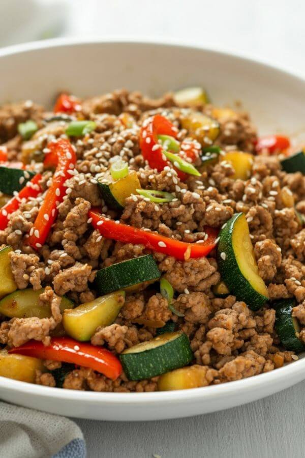 A vibrant and appetizing skillet stir-fry featuring browned ground turkey, diced zucchini, and sliced red bell peppers. The dish should be lightly glazed with soy sauce, garnished with a sprinkle of sesame seeds, and served on a clean white plate for a fresh, colorful presentation. Use natural lighting to emphasize the dish's healthy, wholesome appeal. the background is light