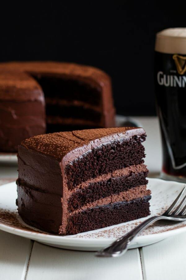 A rich, dark chocolate cake with a glossy frosting, garnished with a sprinkle of cocoa powder. The cake sits on a white wooden table and a fork resting beside the plate.