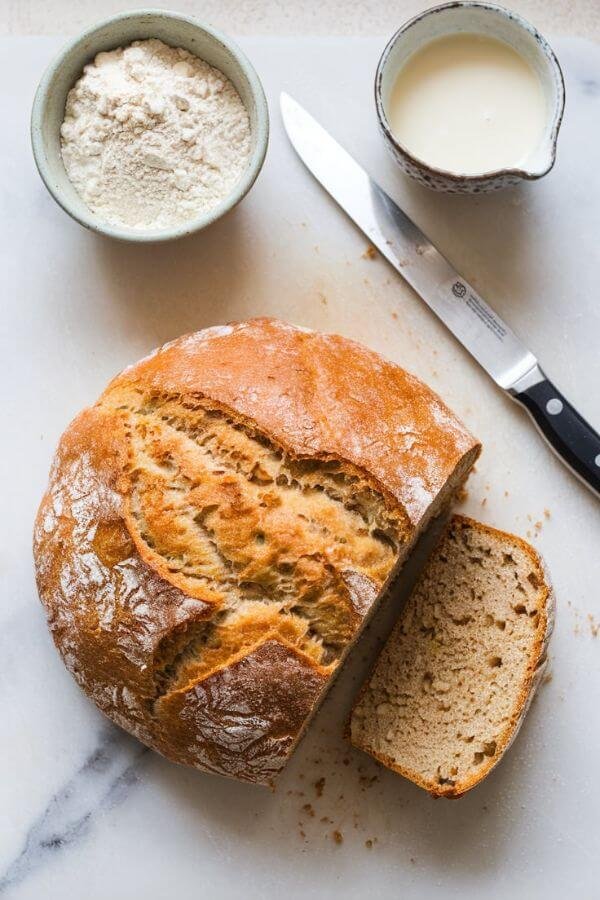 Craft a detailed image of a rustic, round loaf of golden-brown soda bread, resting on a pristine white cutting board. A sharp knife lies beside it, hinting at a recent slice. Nearby, a small bowl brimming with flour and a cup of creamy almond milk sit invitingly.