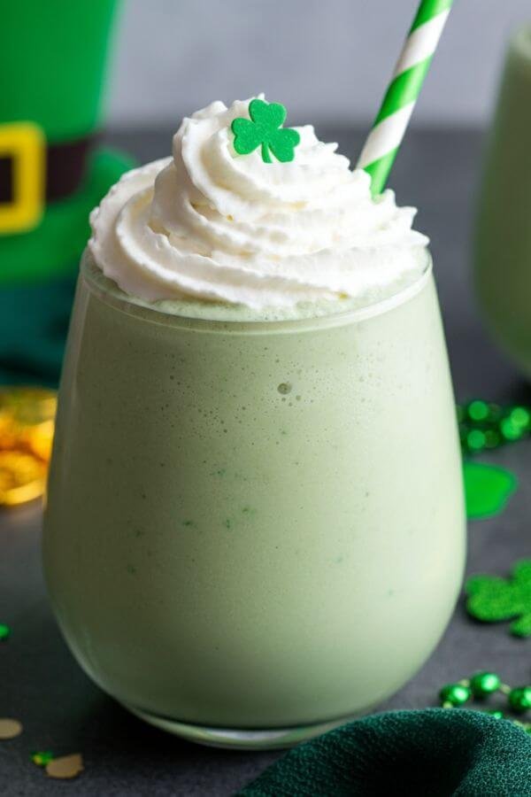 A creamy, green vegan shamrock shake in a clear glass, topped with a dollop of vegan whipped cream and a festive green-and-white striped straw. The background features subtle St. Patrick's Day decor like shamrocks, gold coins, or a touch of green fabric.