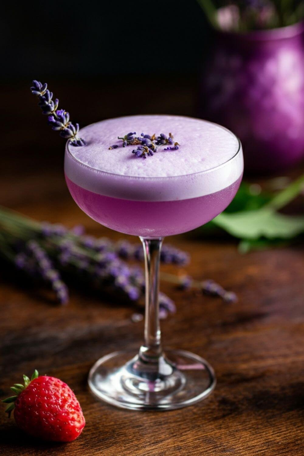 A stylish cocktail glass filled with a pale lavender-hued drink, topped with a frothy layer of foam. A sprig of fresh lavender rests elegantly on the rim, complemented by soft, natural lighting and a light table setting.