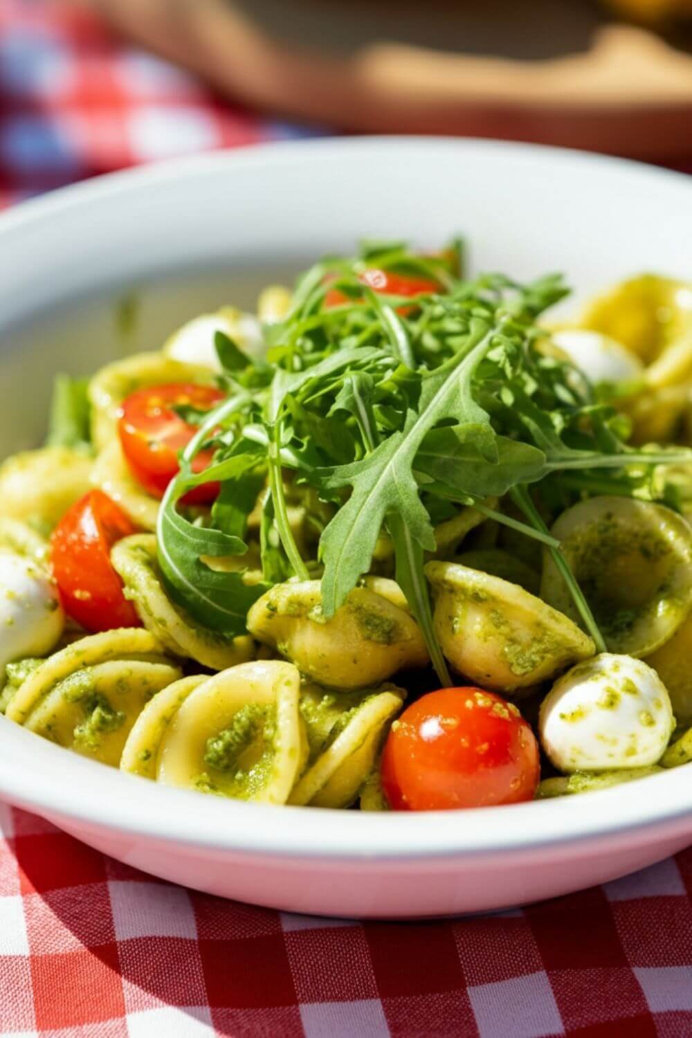 A bright and fresh pesto pasta salad served in a white bowl. The orecchiette is coated in vibrant green pesto, with cherry tomatoes and mozzarella pearls nestled throughout. Garnished with arugula leaves and a sprinkle of Parmesan, the dish sits on a checkered picnic tablecloth in natural light.