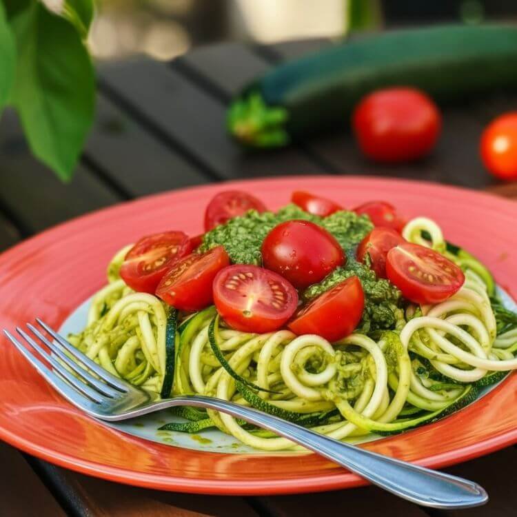 A vibrant plate of zucchini noodles, lightly coated in bright green basil pesto, with juicy cherry tomato halves scattered on top. Finish the look with a sprinkling of parmesan and a fork ready for that first bite. The dish is placed on a rustic wooden table with fresh basil leaves and a wedge of parmesan in the background.